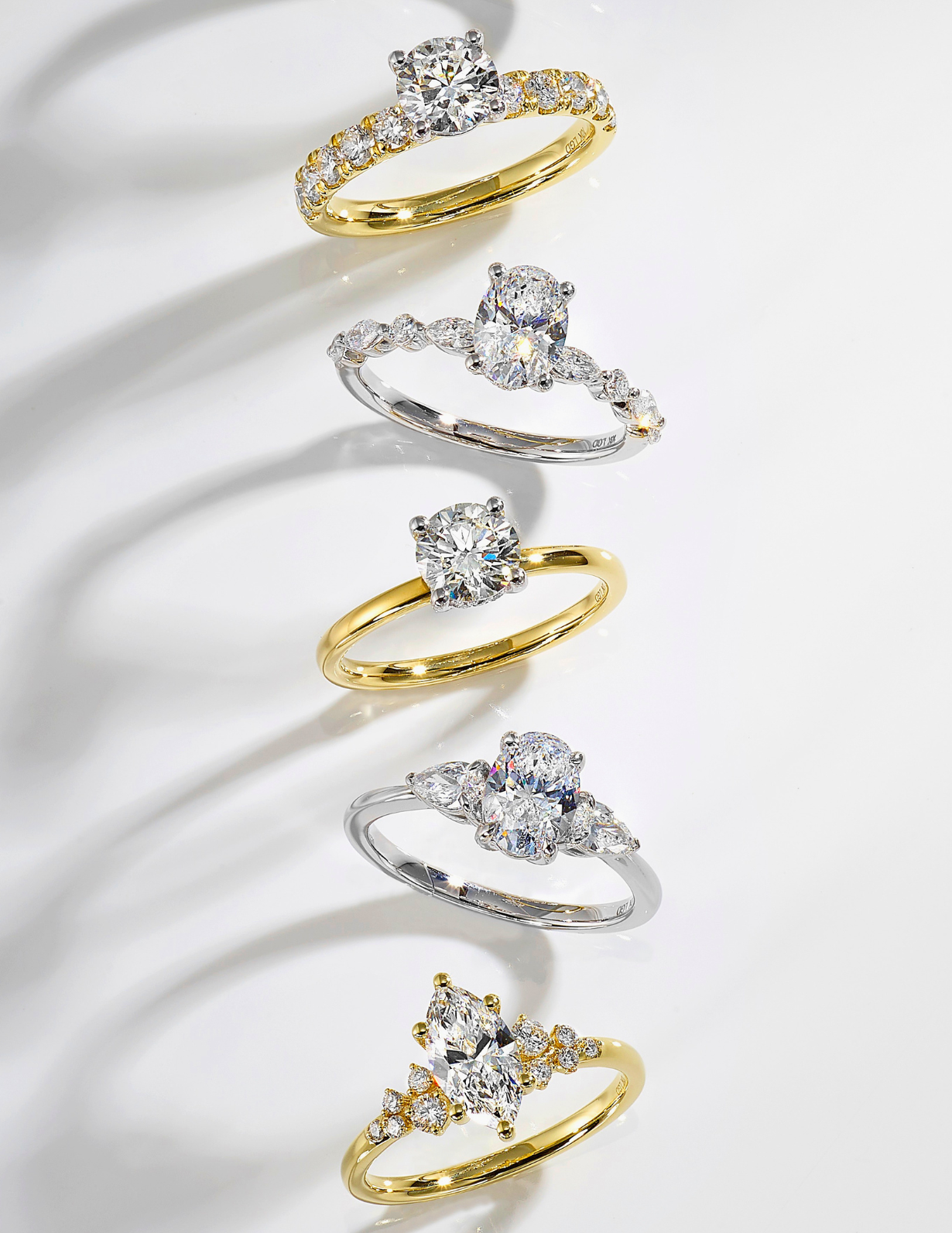 Seven engagement-style rings with round center stones and pavé accents, arranged vertically on a white background. The bands are shown in both yellow gold and silver-tone metals, with some rings featuring small side stones.